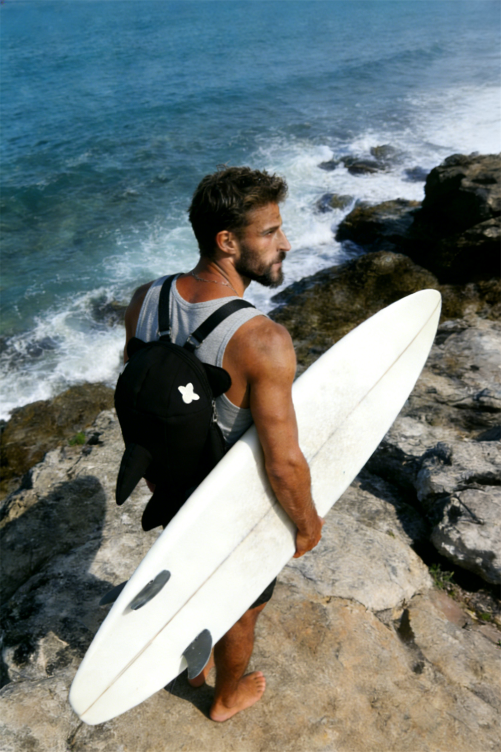 Male surfer on a rocky coast carrying a surfboard and wearing the black SHAKU shark-shaped backpack, showcasing unconventional Japanese youth fashion.