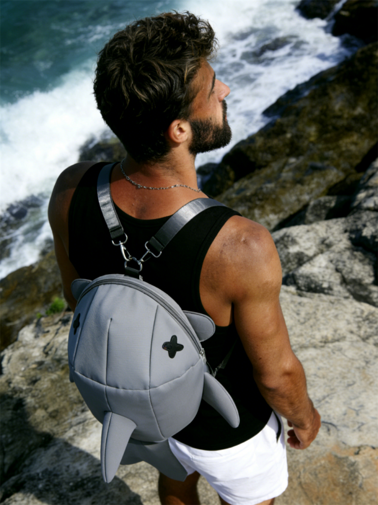 Young male model standing on ocean rocks with a surfboard, wearing the stone grey SHAKU shark backpack with 3D fins and Japanese streetwear design.
